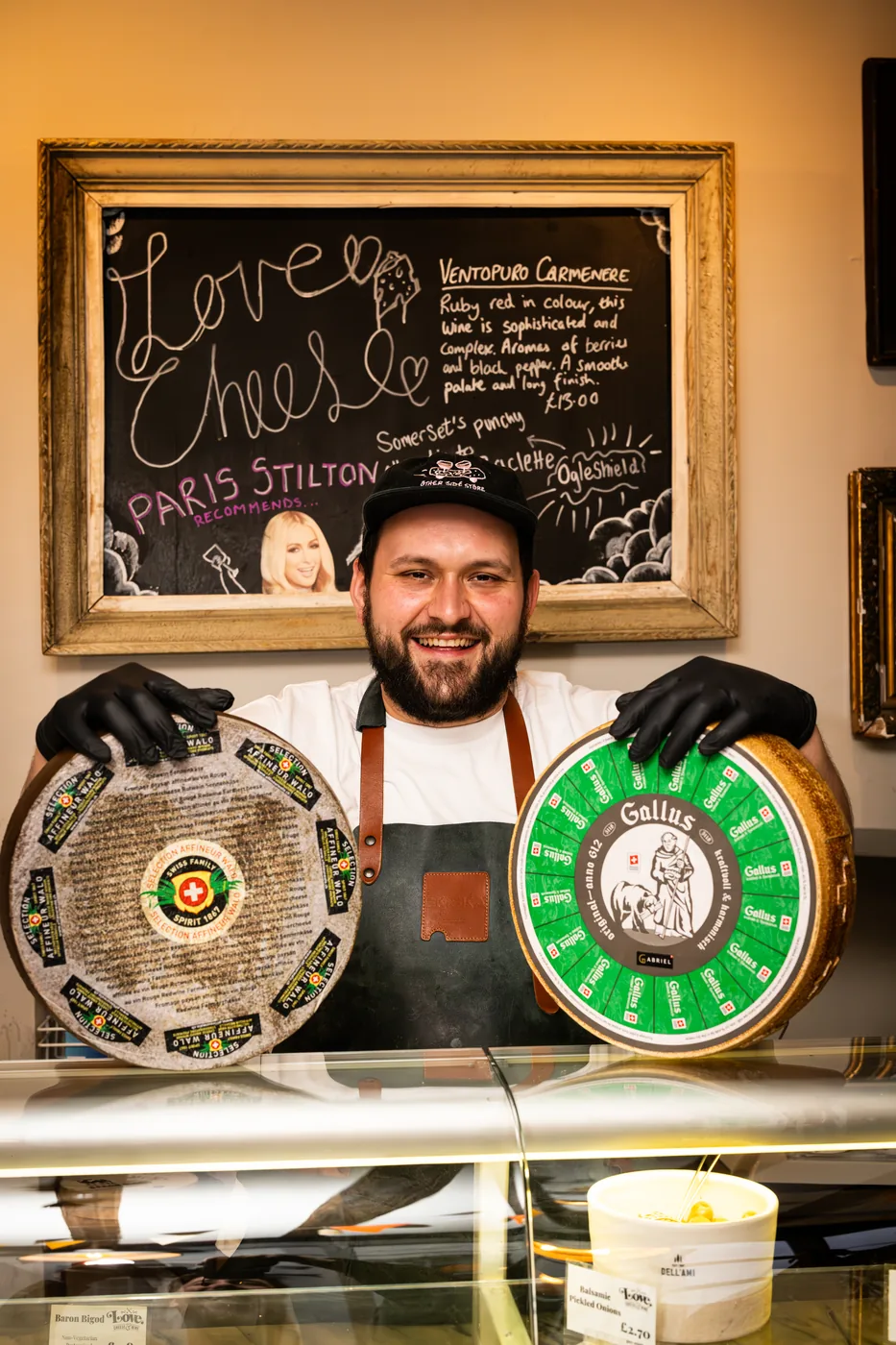 A cheesemonger behind the counter holding two large cheese wheels